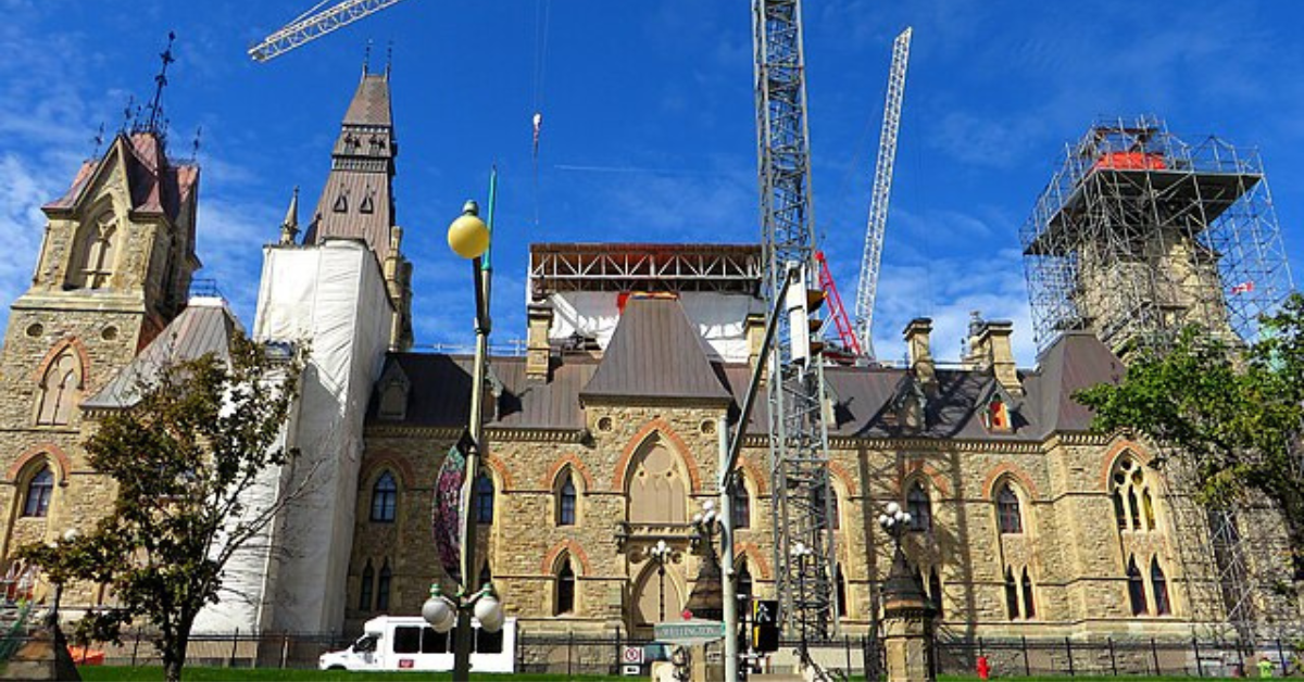 Canadian Parliament's West Block building under heavy restoration, surrounded by scaffolding and construction cranes against a bright blue sky.