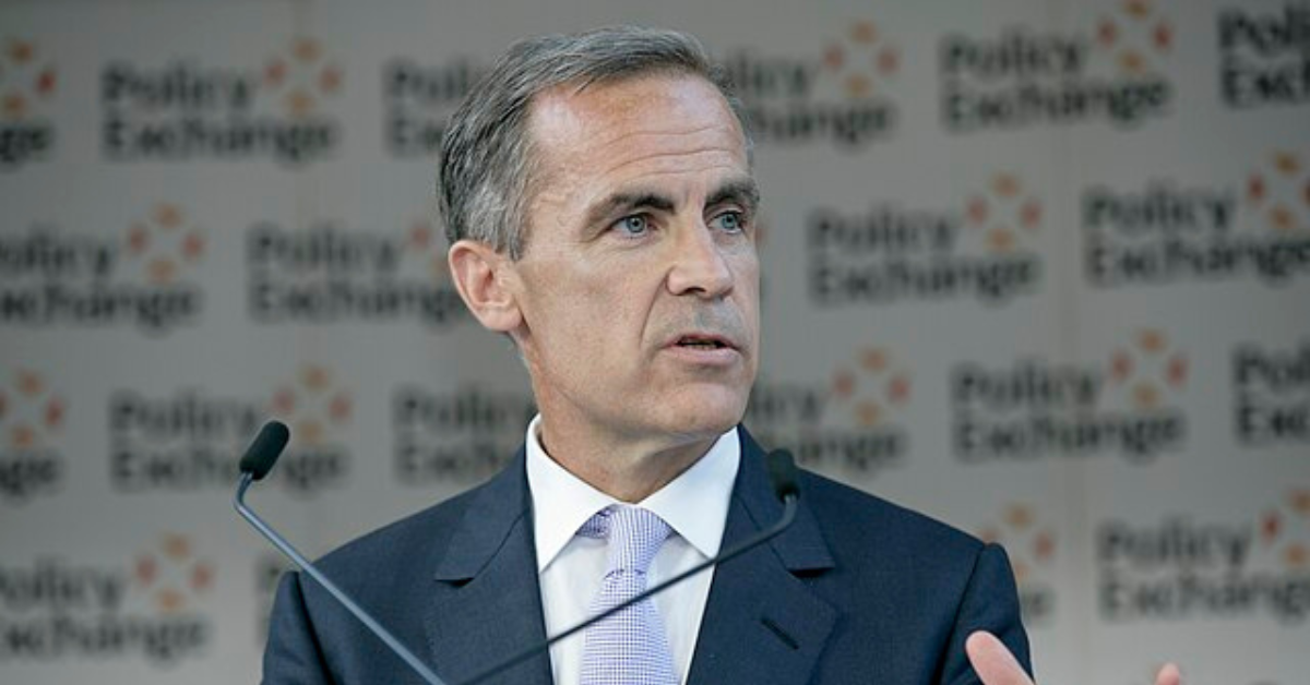 Mark Carney speaking at the Policy Exchange event, standing at a podium in a dark suit and lavender tie, with two microphones in front of him.